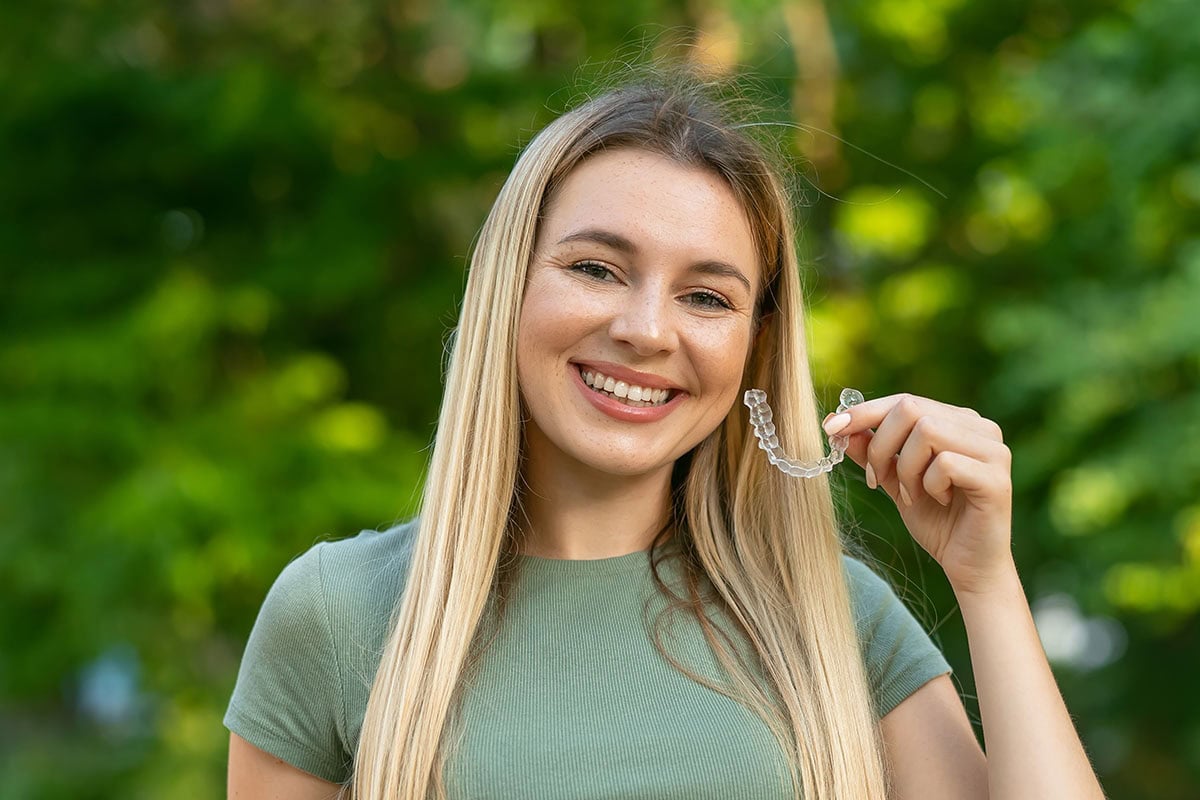 Lächelnde blonde Frau im Freien hält einen durchsichtigen Aligner.